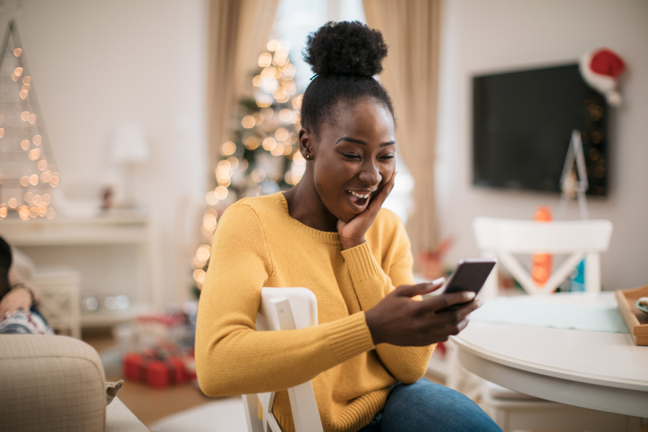 Happy young African American woman using a smart phone, staying in touch with her family and friends and wishing them a Marry Christmas and a happy new year